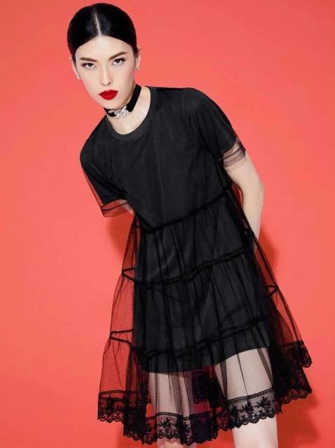 Woman in a black tiered dress with lace trim against a bright red background in a studio setting, fashion editorial shot, portrait orientation.