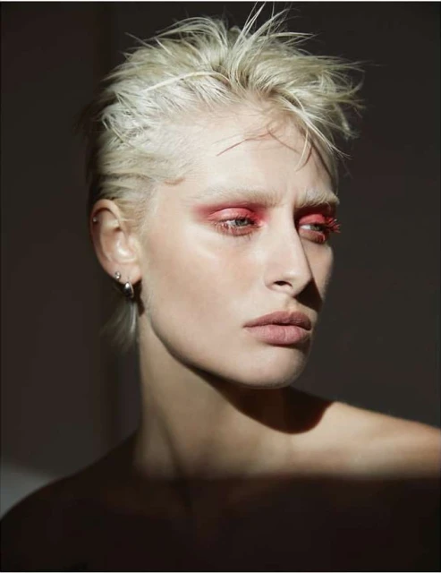 Close-up portrait of an adult woman with short blonde hair and bold pink eyeshadow looking to the side, against a shadowy background.