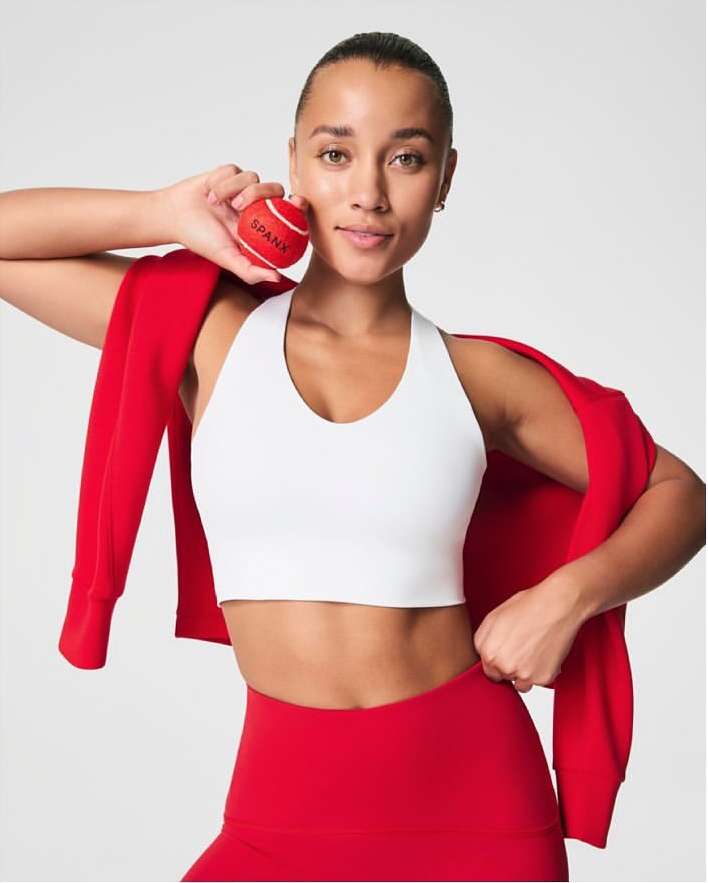 Athletic woman in a white sports bra and red leggings smiles while holding a red Spanx-branded tennis ball in a brightly lit setting.