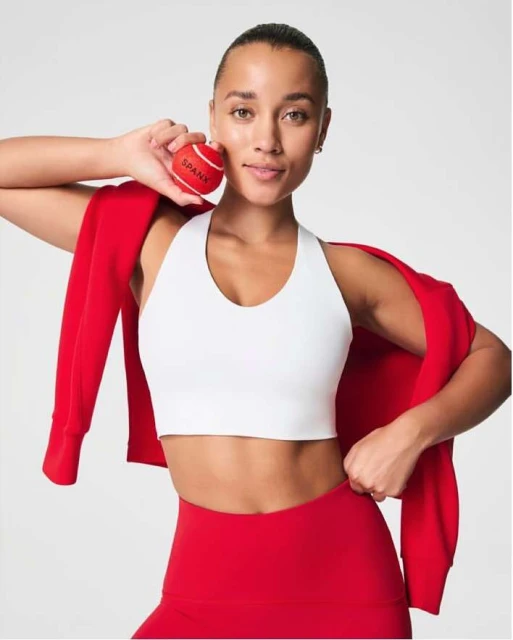 Athletic woman in a white sports bra and red leggings smiles while holding a red Spanx-branded tennis ball in a brightly lit setting.