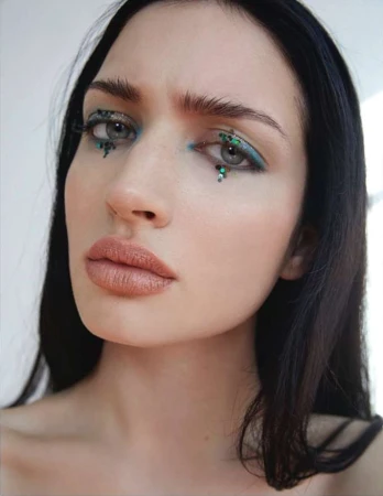 Close-up shot of an adult woman with creative makeup, including blue eyeliner and green glitter under her eyes, set against a white background.