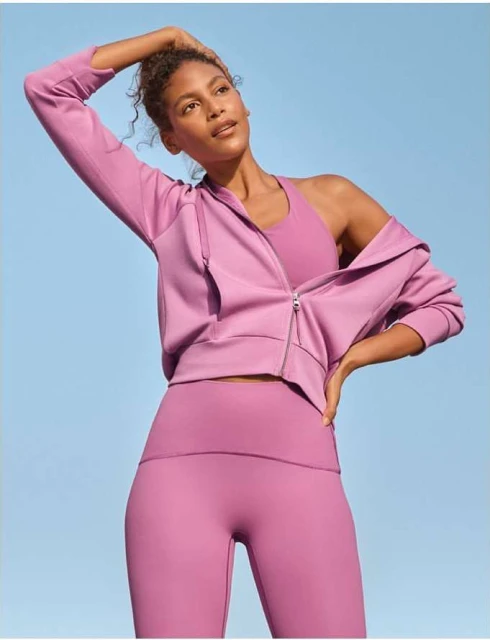 An adult female in workout clothes, including leggings, a sports bra, and a zip-up jacket, poses against a blue sky backdrop in a fitness-style shot.