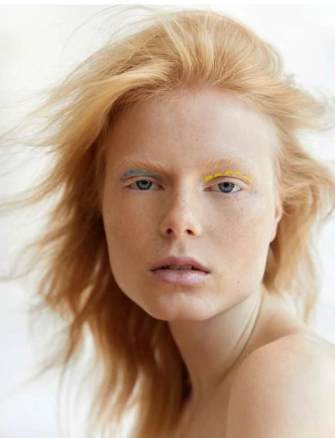 Close-up portrait of a young adult woman with pale skin, red hair blowing in the wind, and blue and yellow eyeliner.