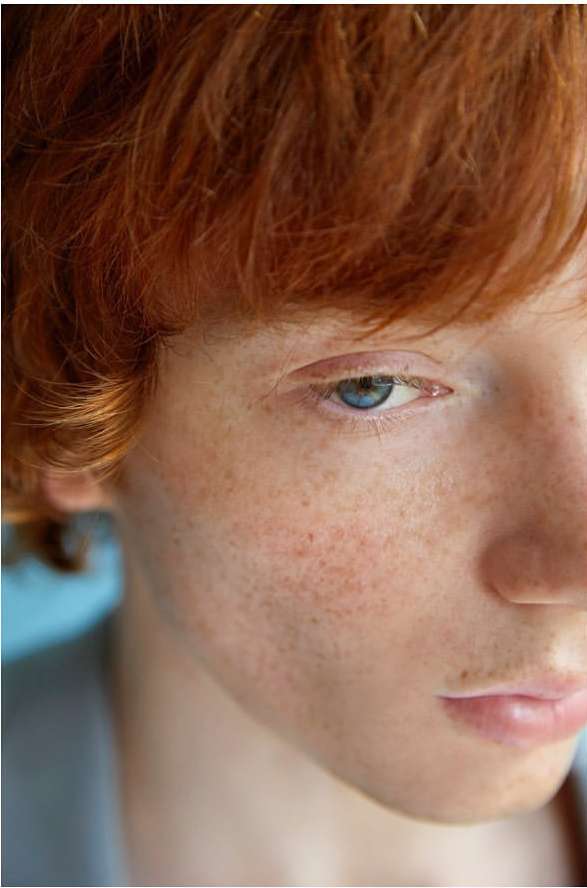 Close-up portrait of a young person with red hair, blue eyes, and visible freckles captured by photographer Kimber Capriotti.