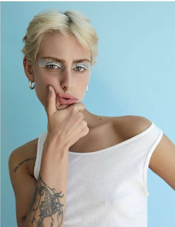 Close-up of a fair-skinned woman with short blonde hair, glitter eyeshadow, and a white tank top against a light blue backdrop.