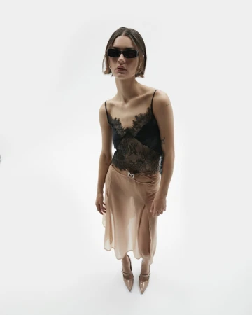 Woman in dark sunglasses, black lace camisole, and sheer nude skirt posing against a seamless white studio background.