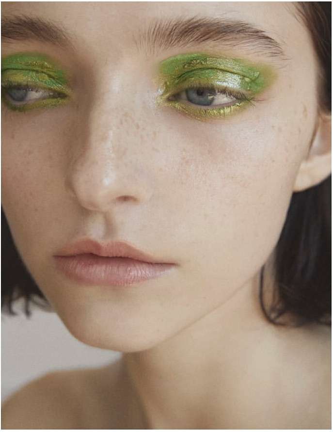 Close-up portrait of a young adult woman with vibrant green eye makeup, fair skin, and a subtle, contemplative facial expression.