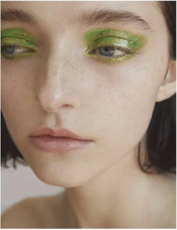 Close-up portrait of a young adult woman with vibrant green eye makeup, fair skin, and a subtle, contemplative facial expression.
