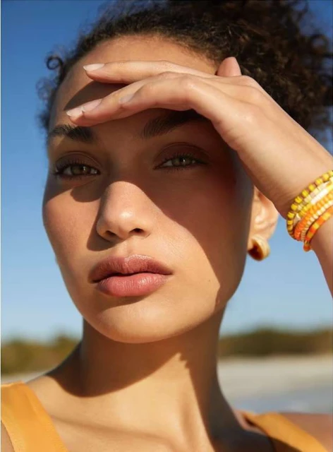 Close-up portrait of an adult female model shielding her eyes from the sun, wearing orange beaded bracelets, with a beach backdrop.