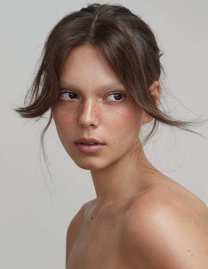 Close-up portrait of a woman with freckles, brown hair, and side bangs against a neutral studio backdrop