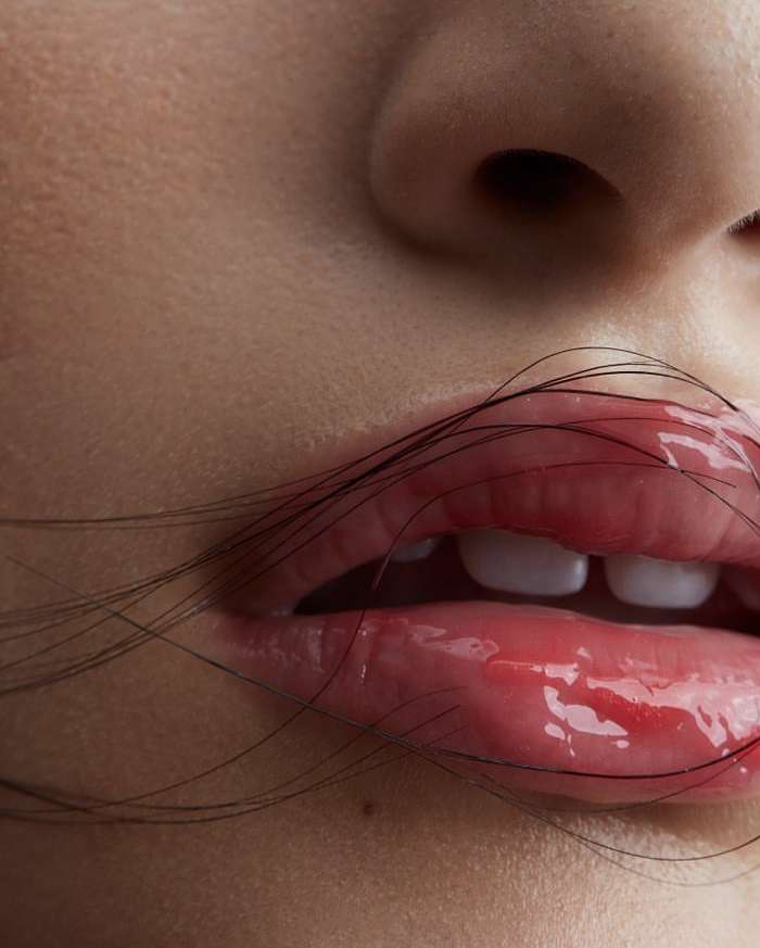 Close-up of a person's face featuring glossy pink lips partially covered with thin strands of dark hair against a neutral background.