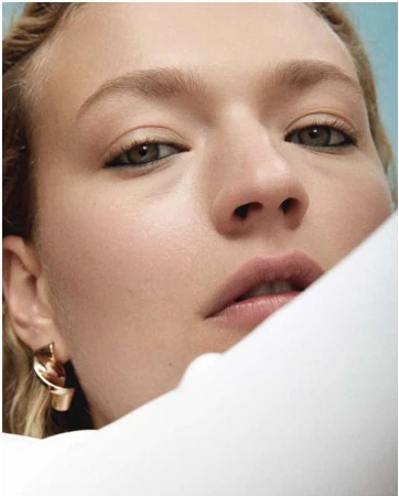 Close-up portrait of a woman with dark eyeliner and a metallic twist earring against a blue background