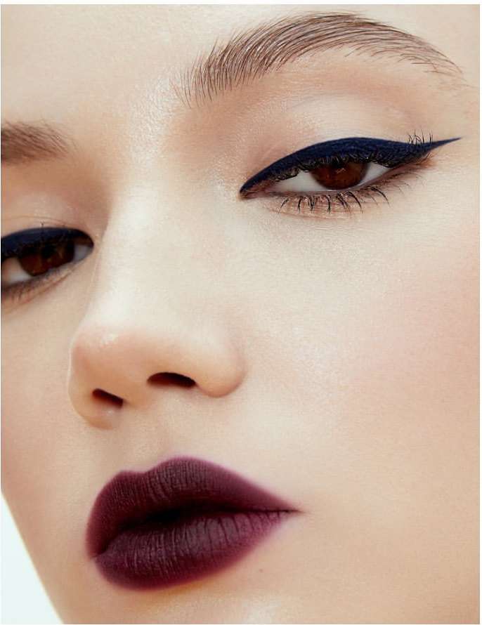 Close-up of a woman's face highlighting her makeup, including bold winged eyeliner and dark berry lipstick against a light background.