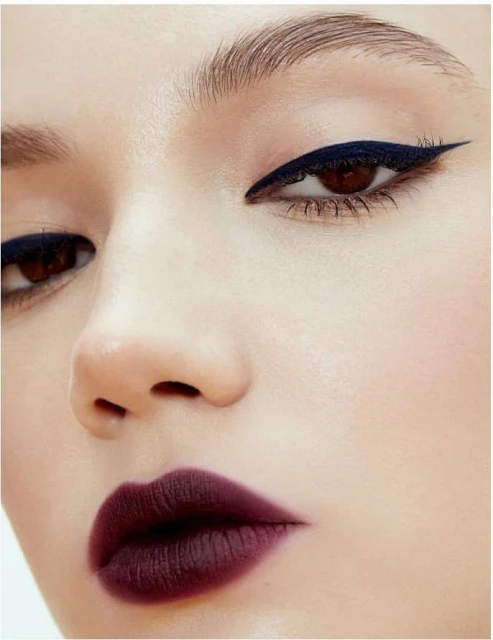 Close-up of a woman's face highlighting her makeup, including bold winged eyeliner and dark berry lipstick against a light background.