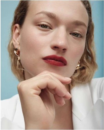 Close-up beauty portrait of a woman with bold red lipstick and gold earrings, wearing a white blazer against a soft blue background.