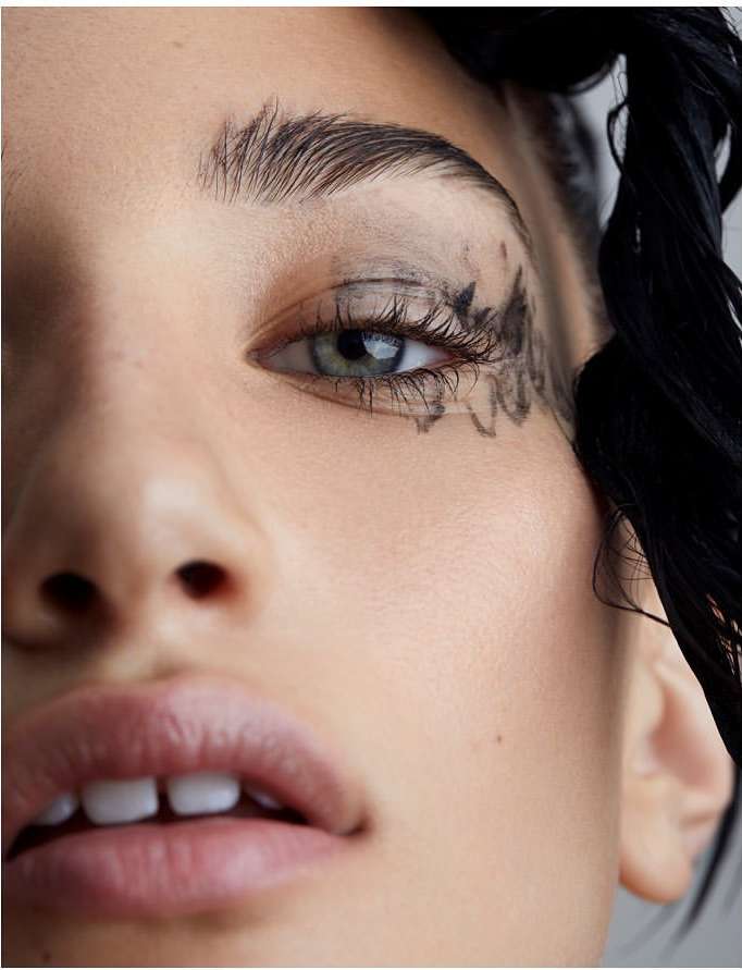 Close-up portrait of a woman with artistic dark eyeliner and dark eyebrow makeup. The woman has blue-green eyes, fair skin, and dark hair.