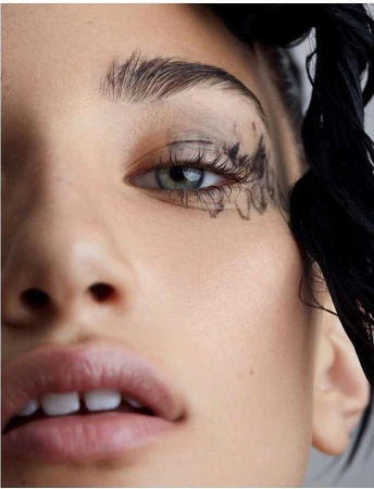 Close-up portrait of a woman with artistic dark eyeliner and dark eyebrow makeup. The woman has blue-green eyes, fair skin, and dark hair.