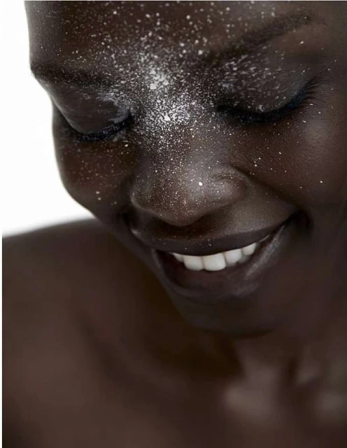 Close-up portrait of a smiling African American woman with white powder sprinkled on her face, set against a bright white backdrop.