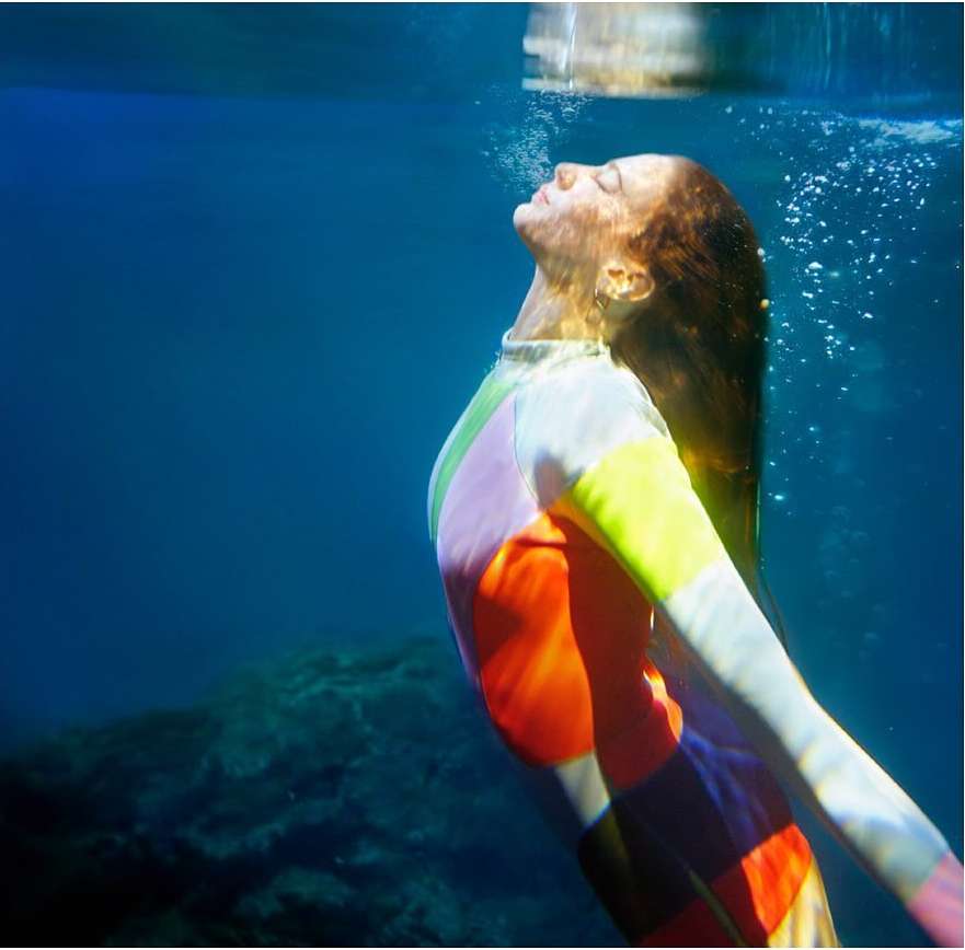 Underwater shot of a woman in a colourful wetsuit, gazing upwards with bubbles around her face in a dark-blue ocean environment.