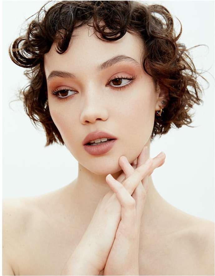 Close-up portrait of a young woman with curly dark hair, flawless skin, and neutral makeup posing against a bright white background.