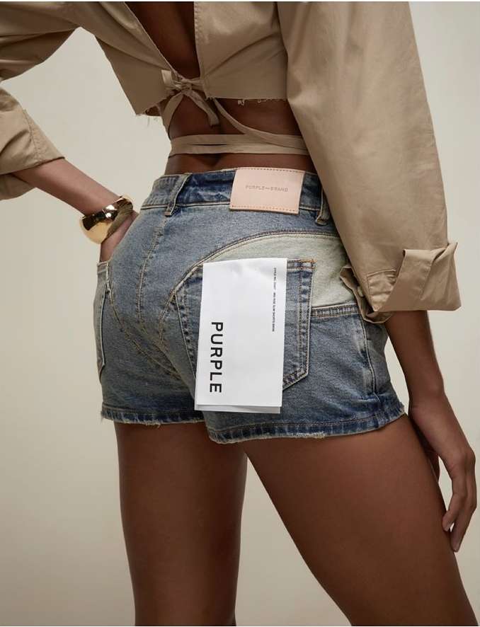Back view of a slender tan-skinned person wearing distressed denim shorts, gold bracelet, and a beige crop top in a studio setting.