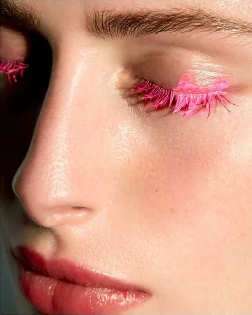 Close-up of a woman's face, highlighting pink eyelashes and glossy lips in a beauty-focused shot, showcasing unique makeup and skin texture.