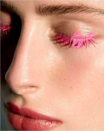 Close-up of a woman's face, highlighting pink eyelashes and glossy lips in a beauty-focused shot, showcasing unique makeup and skin texture.