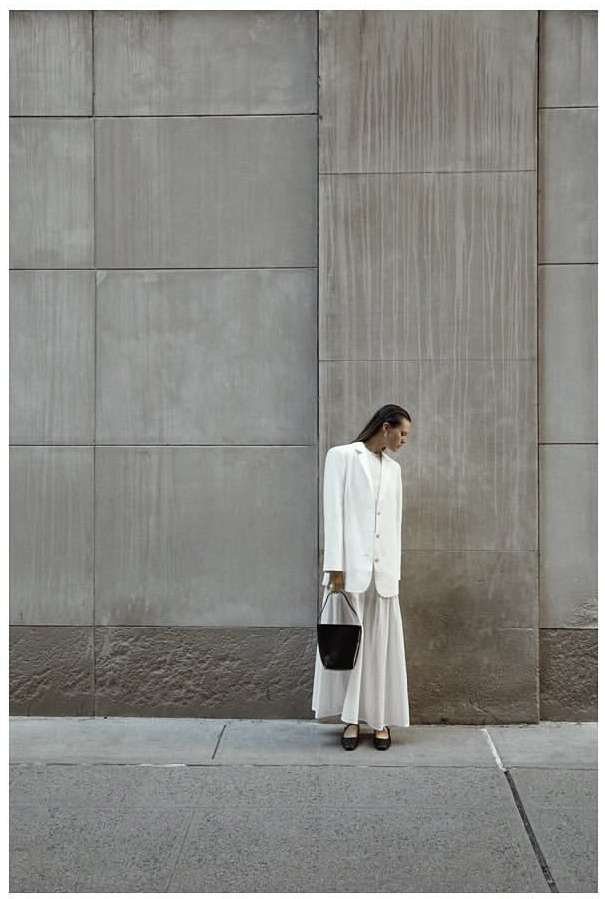 A woman in a white suit and skirt carries a black bucket bag while standing on a sidewalk against a textured grey wall.