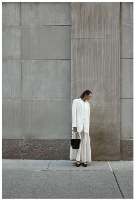 A woman in a white suit and skirt carries a black bucket bag while standing on a sidewalk against a textured grey wall.