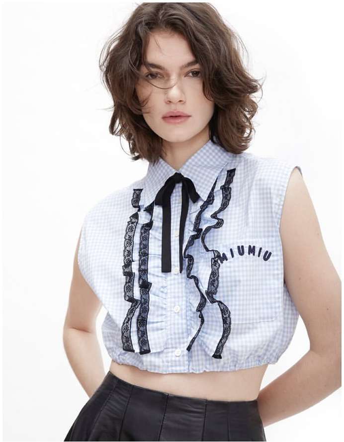 Young adult woman wearing a blue gingham crop top with ruffles and a black ribbon poses in a brightly lit, white studio environment.