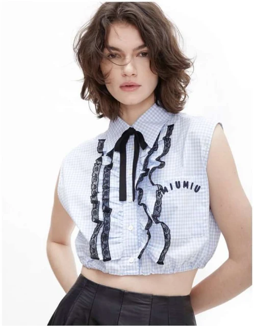 Young adult woman wearing a blue gingham crop top with ruffles and a black ribbon poses in a brightly lit, white studio environment.