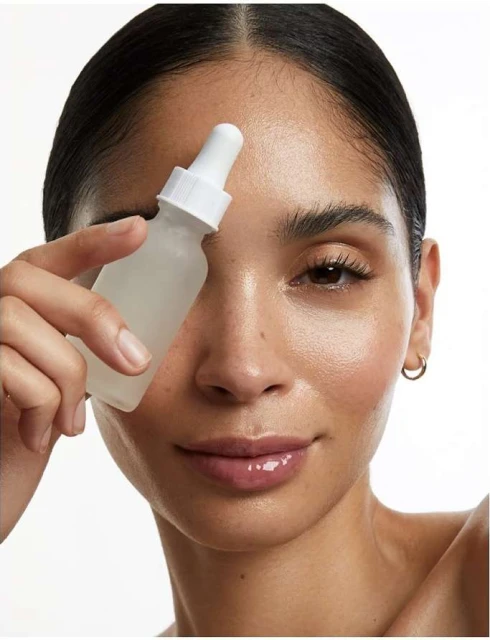 Close-up of a woman holding a translucent bottle with a dropper, skincare concept, indoor photo studio shot against a plain white backdrop.