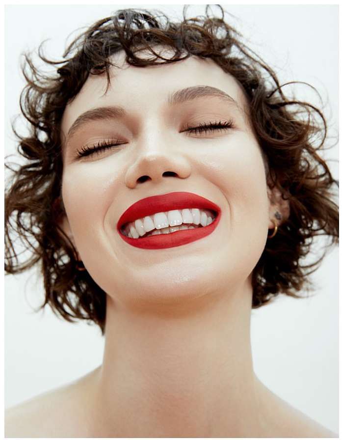 Close-up portrait of a young adult female with curly brown hair, eyes closed, white teeth, and bright red lipstick against a white backdrop.