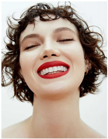 Close-up portrait of a young adult female with curly brown hair, eyes closed, white teeth, and bright red lipstick against a white backdrop.