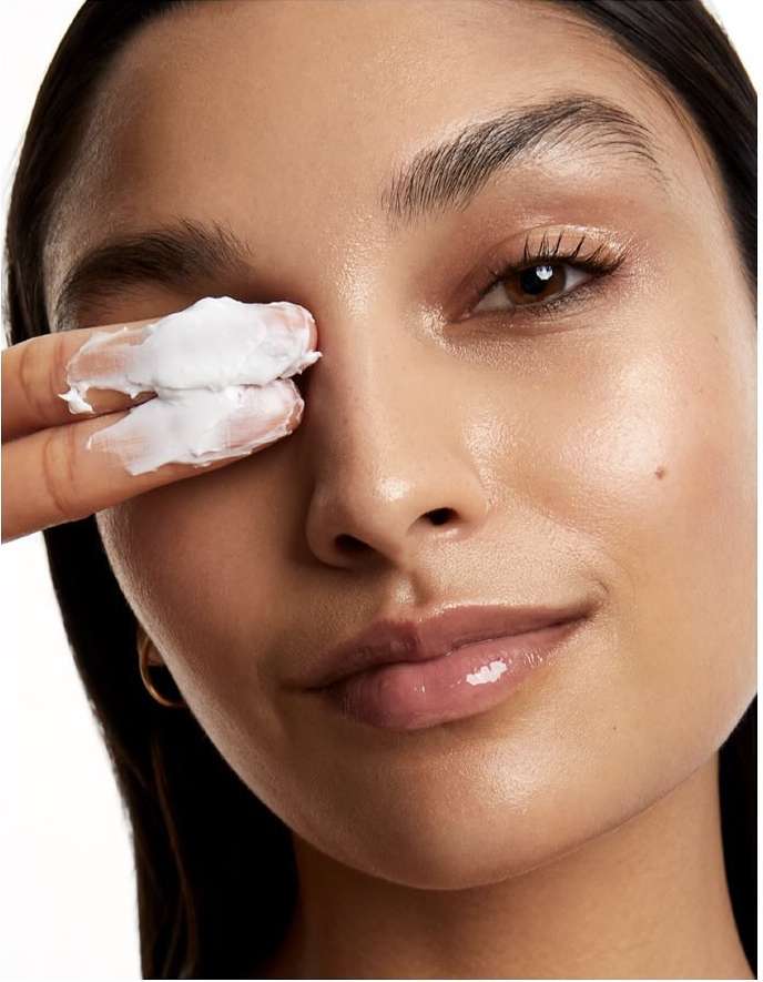 Close-up of a woman's face as she applies white cream to her eye area with her fingers, showcasing skincare and beauty.