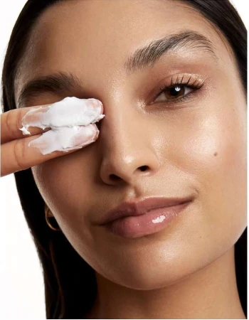 Close-up of a woman's face as she applies white cream to her eye area with her fingers, showcasing skincare and beauty.
