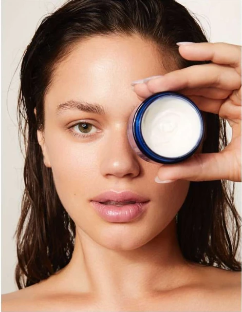 Adult female presents a small container of cosmetic face cream, portrait shot in photo studio setting, demonstrating beauty product.