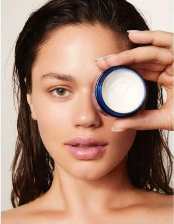Adult female presents a small container of cosmetic face cream, portrait shot in photo studio setting, demonstrating beauty product.