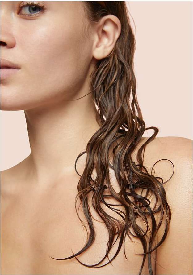 Close-up of a woman with fair skin and shoulder-length, wet, wavy brown hair against a peach background, showcasing a beauty aesthetic.