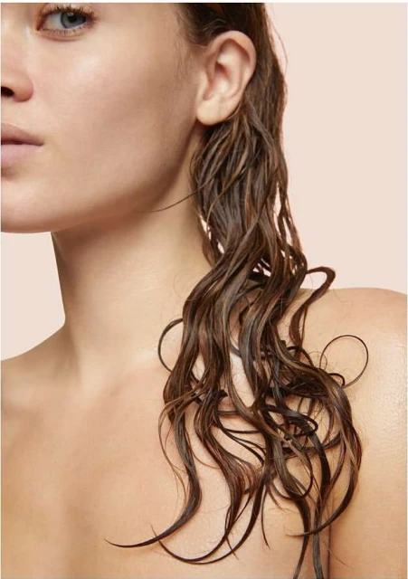 Close-up of a woman with fair skin and shoulder-length, wet, wavy brown hair against a peach background, showcasing a beauty aesthetic.
