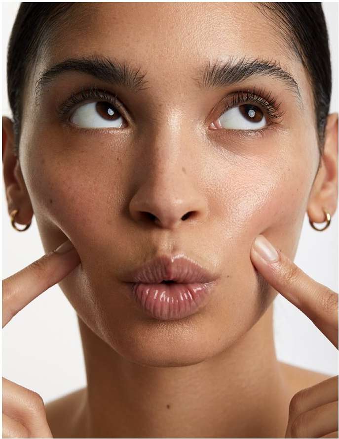 Close-up of a young woman with fair skin, puckered lips, and gold hoop earrings, playfully touching her cheeks with her index fingers.