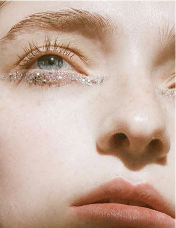 Close-up beauty portrait of a woman with pale skin, blue eyes, and sparkling silver glitter applied in a sharp line beneath her lower eyelids.