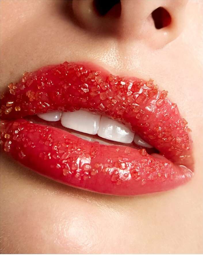 Close-up of a woman's lips covered in red sugar crystals; showing teeth. The image is a beauty shot with a focus on lip detail.