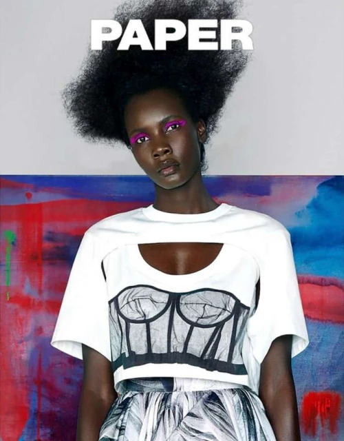Editorial fashion shot of a Black female model with an afro hairstyle and makeup, wearing a cutout top and patterned skirt against a colourful backdrop.