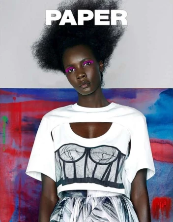 Editorial fashion shot of a Black female model with an afro hairstyle and makeup, wearing a cutout top and patterned skirt against a colourful backdrop.