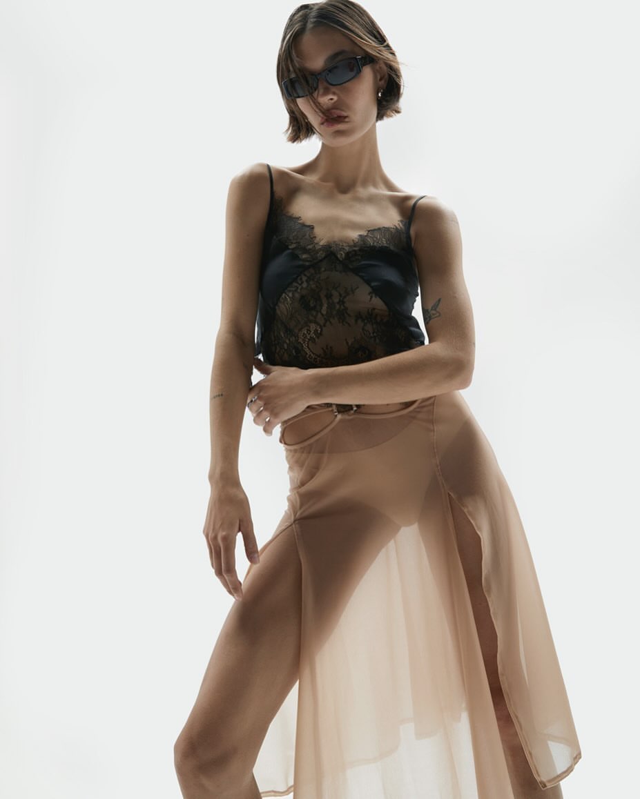 A woman with short brown hair poses in sunglasses, a black lace top, and a tan sheer skirt against a bright white backdrop in a studio setting.