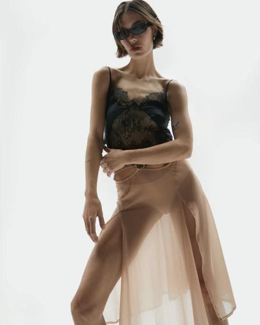 A woman with short brown hair poses in sunglasses, a black lace top, and a tan sheer skirt against a bright white backdrop in a studio setting.