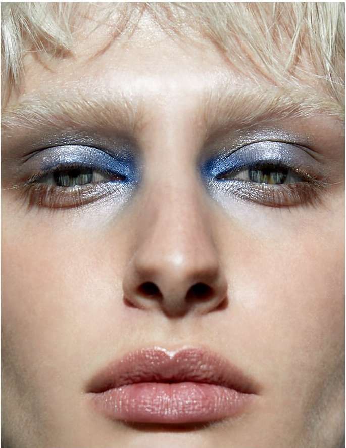 Close-up portrait of a woman with blue eyeshadow and pale skin, highlighting makeup and facial features in a neutral setting.