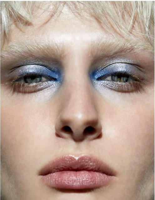 Close-up portrait of a woman with blue eyeshadow and pale skin, highlighting makeup and facial features in a neutral setting.