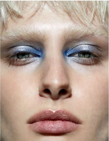 Close-up portrait of a woman with blue eyeshadow and pale skin, highlighting makeup and facial features in a neutral setting.
